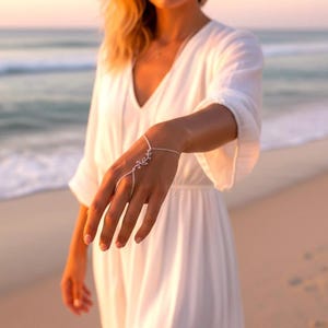 Puede incluir: Una delicada cadena de mano plateada con un diseño de hojas. La joya se conecta a un anillo y se envuelve alrededor de la muñeca. El fondo presenta una escena de playa con el océano y una persona con un vestido blanco.