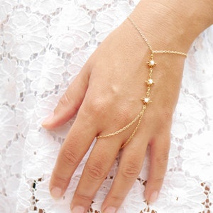Puede incluir: Un brazalete de cadena dorada con un diseño delicado que presenta tres dijes en forma de estrella con piedras blancas. El brazalete está conectado a un anillo que se puede usar en el dedo.