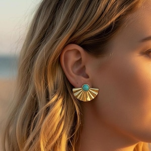 Peut inclure: Boucles d'oreilles dorées en forme d'éventail avec une pierre centrale turquoise. Les boucles d'oreilles ont un motif de rayons de soleil et sont portées sur l'oreille. L'image montre un gros plan des boucles d'oreilles.