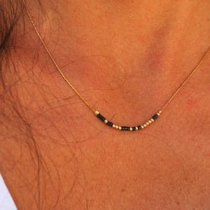 Op de afbeelding: Een delicate gouden ketting met een reeks kleine zwarte en gouden kralen. De ketting wordt om de nek gedragen, waarbij de kralen op de huid rusten. De ketting is een eenvoudig, elegant sieraad.
