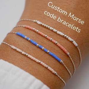 May include: Four delicate bracelets with beaded Morse code designs. The bracelets feature silver chains and beads in various colors, including pink, blue, and white. The text "Custom Morse code bracelets" is visible in the image.