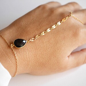 May include: A gold chain bracelet with a black teardrop-shaped gemstone pendant. The bracelet is designed to be worn on the wrist and has a chain that extends up the arm to the finger.