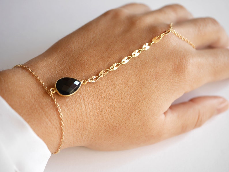 May include: A gold chain bracelet with a black teardrop-shaped gemstone pendant. The bracelet is designed to be worn on the wrist and has a chain that extends up the arm to the finger.
