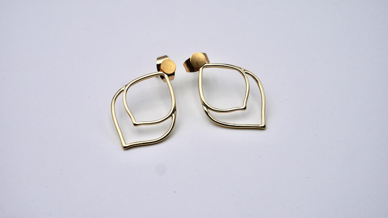 May include: A pair of gold-colored leaf-shaped earrings. Each earring features a double-layered design with a post backing. The earrings are displayed on a white surface, showcasing their minimalist and elegant style.