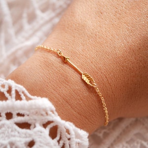 Könnte beinhalten: Ein zartes, goldfarbenes Armband mit einem Pfeildesign. Der Pfeil ist der Mittelpunkt, mit detaillierten Federn. Das Armband befindet sich an einer feinen Kette und wird am Handgelenk getragen. Der Schmuck befindet sich vor einem weichen, neutralen Hintergrund.