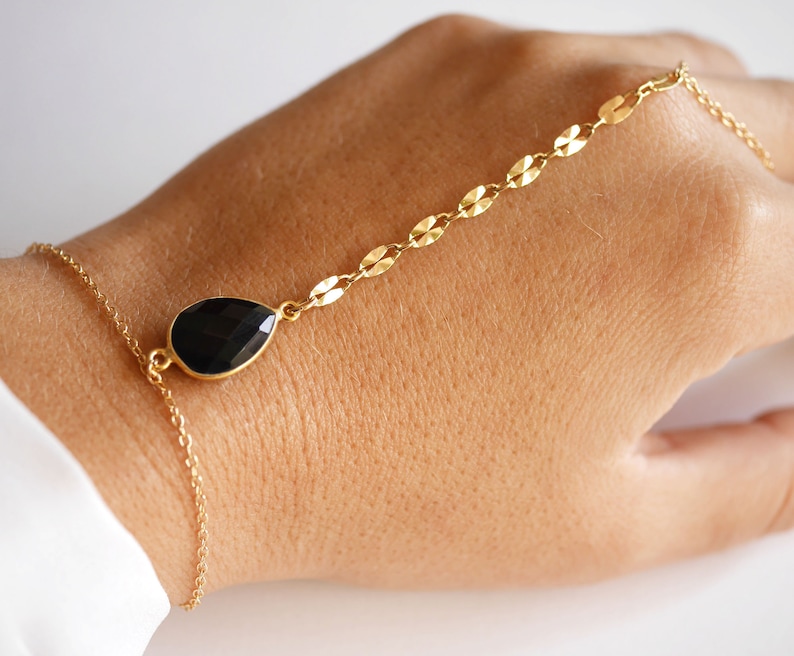 May include: A gold chain bracelet with a black teardrop-shaped gemstone. The bracelet is designed to be worn around the wrist and up the arm.