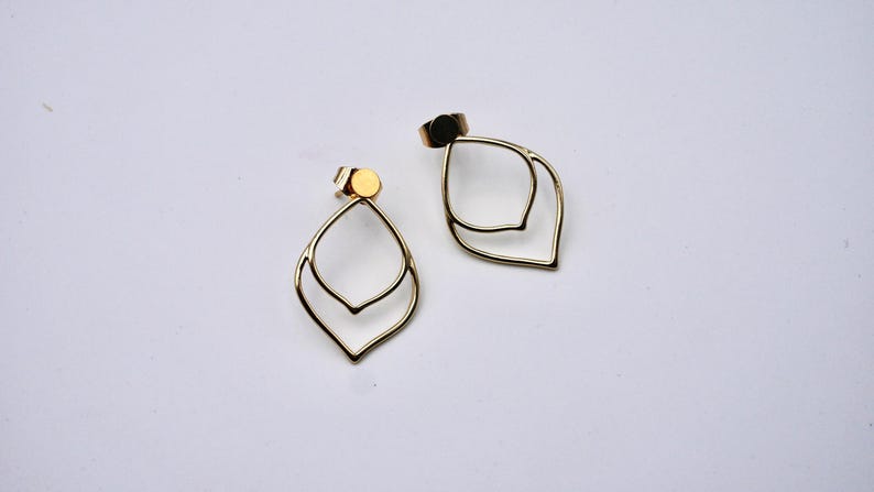 May include: A pair of gold-colored, open-frame earrings. Each earring features a double-layered, teardrop-shaped design. The earrings are displayed on a white surface, showcasing their minimalist and elegant style. The earring posts are visible.