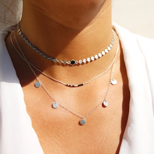 Pode incluir: Um colar de prata em camadas com uma camada superior de estilo choker composta por pequenos discos de prata e uma camada inferior com discos de prata pendurados.
