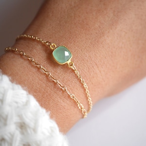 May include: A delicate gold chain bracelet with a light green gemstone pendant. The pendant is a square shape with rounded edges.