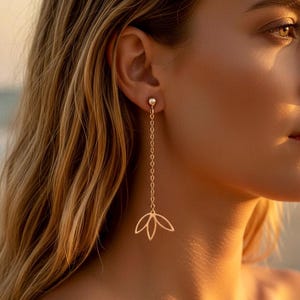Peut inclure: Une boucle d'oreille dorée avec une chaîne et un motif de fleur de lotus. La boucle d'oreille est suspendue à l'oreille. L'arrière-plan est flou, avec des tons chauds.