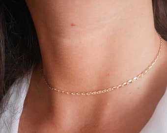 May include: A delicate gold chain necklace with a simple, flat link design. The necklace is worn close to the neck, creating a minimalist and elegant look.