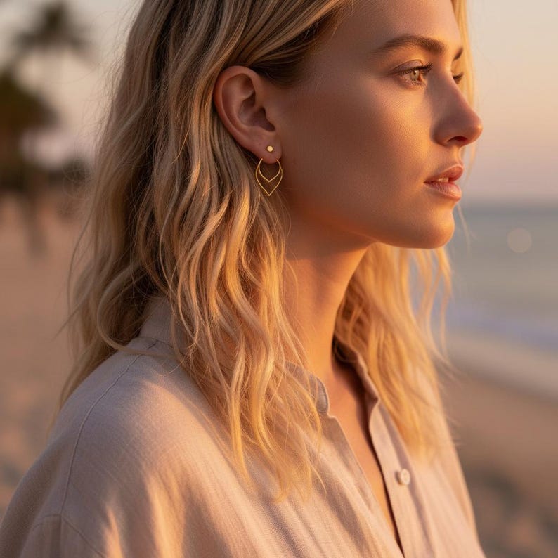 May include: A pair of gold earrings. The earrings feature a small stud and a dangling teardrop design. The earrings are worn by a person with blonde hair, and the background is a blurred view of a beach at sunset.