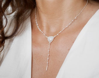 Può includere: Una collana d'argento con un ciondolo di pietra bianca e una lunga catena delicata. La catena è adornata con piccole perle bianche.