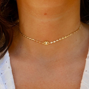 Può includere: Una collana a catena dorata con un piccolo ciondolo a forma di occhio con una pietra trasparente al centro. La collana è sul collo di una persona.
