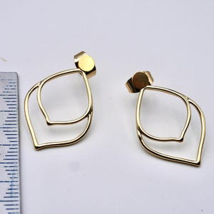May include: A pair of gold-colored, layered leaf-shaped stud earrings. The earrings have a post backing and are displayed on a white surface. A ruler is placed next to the earrings.