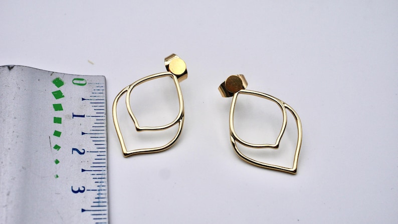 May include: A pair of gold-colored, layered leaf-shaped stud earrings. The earrings have a post backing and are displayed on a white surface. A ruler is placed next to the earrings.