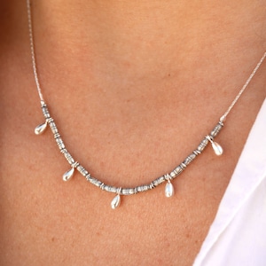 Puede incluir: Un collar de plata con una cadena delicada y pequeños colgantes en forma de lágrima. Los colgantes están espaciados uniformemente a lo largo de la cadena y cuelgan ligeramente.