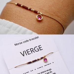 May include: Delicate gold-coloured bracelet with dark red beads and a small, round pink gemstone pendant. The bracelet is a Morse code design. The word "VIERGE" is printed on the accompanying card, which also has Morse code symbols.