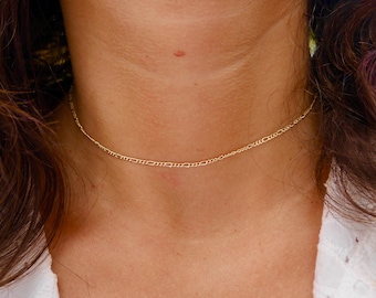 May include: A delicate gold chain necklace, worn as a choker, rests on a person's neck. The chain is thin and has a simple, elegant design.
