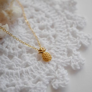 Peut inclure: Un collier avec un pendentif en forme d'ananas en or sur une surface en crochet blanc.