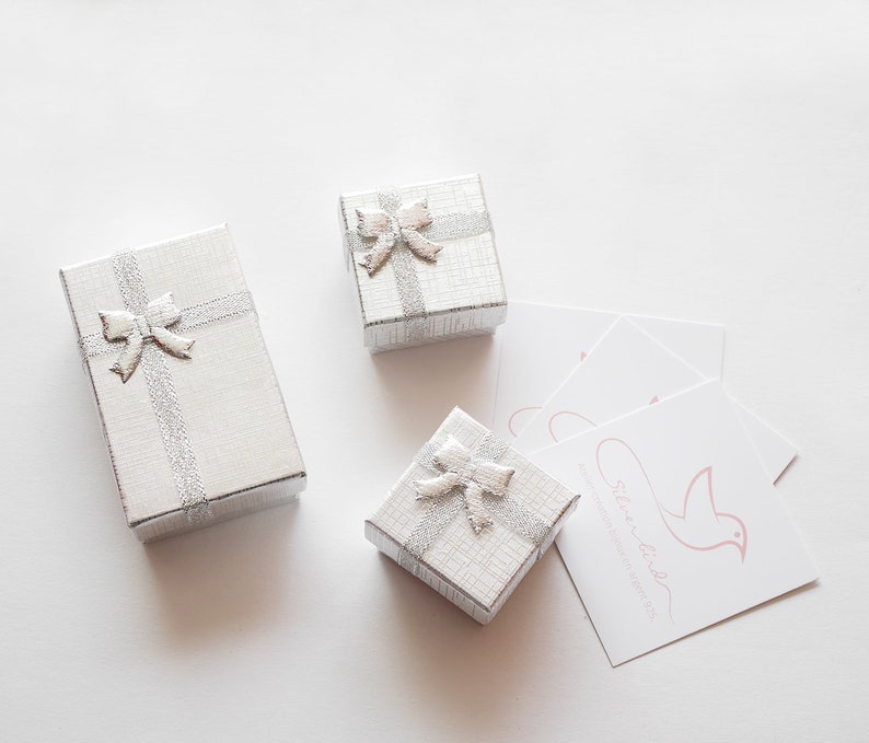 May include: Three silver gift boxes with matching silver bows and ribbons. The boxes are rectangular and have a textured pattern. Several white cards with a pink bird logo and text are also present.