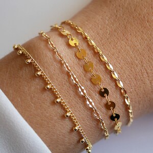 May include: Four gold bracelets stacked on a wrist. The top bracelet has a delicate chain with small, round gold charms. The second bracelet has a thicker chain with larger, round gold charms. The third bracelet has a delicate chain with small, round gold charms. The bottom bracelet has a delicate chain with small, round gold charms.