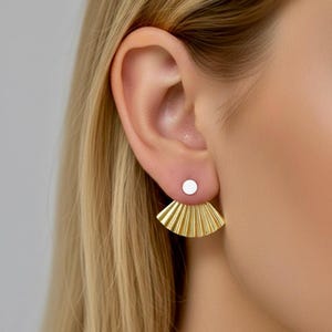 Peut inclure: Gros plan sur une paire de boucles d'oreilles dorées. Les boucles d'oreilles sont composées d'un petit clou circulaire blanc et d'un dos texturé en forme d'éventail. Elles sont portées sur une oreille, avec des cheveux blonds visibles.