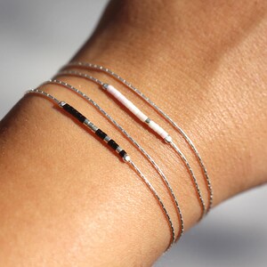 Pode incluir: Um conjunto de quatro pulseiras delicadas de prata com contas de diferentes cores. As pulseiras est&atilde;o sobrepostas no pulso de uma pessoa.