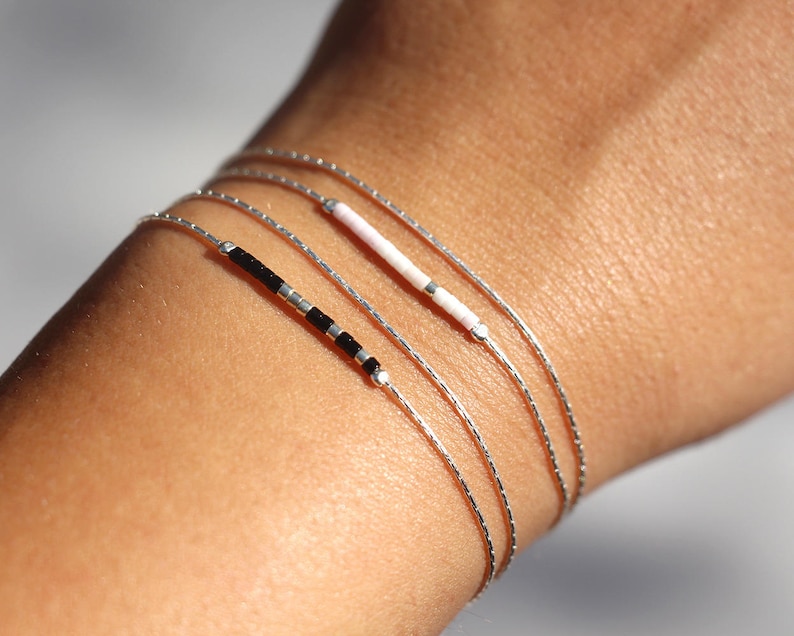 Pode incluir: Um conjunto de quatro pulseiras delicadas de prata com contas de diferentes cores. As pulseiras est&atilde;o sobrepostas no pulso de uma pessoa.