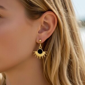 Könnte beinhalten: Goldfarbene Ohrringe mit Sonnenstrahlen-Design. Jeder Ohrring hat eine schwarze kreisförmige Mitte und strahlenförmige Linien, die ein sonnenähnliches Aussehen erzeugen. Die Ohrringe sind hängend und haben einen Steckerverschluss.