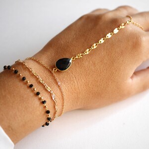 May include: A gold chain bracelet with a black gemstone pendant, layered with two other gold chains and a black beaded chain.