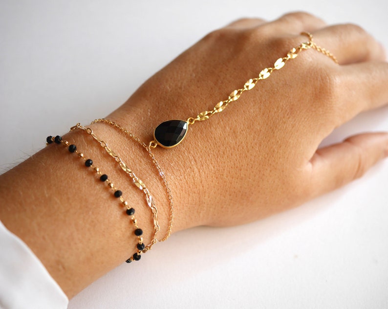 May include: A gold chain bracelet with a black gemstone pendant, layered with two other gold chains and a black beaded chain.