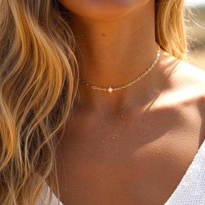 Peut inclure: Un délicat collier ras du cou de couleur or avec un petit pendentif en forme d'éclat d'étoile. Le collier est porté autour du cou, et la chaîne a un design subtil et texturé. L'arrière-plan est flou, suggérant un cadre extérieur.