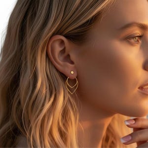 May include: Gold earrings featuring a small stud and a larger, leaf-shaped design. The earrings are worn on a person's ear, with the focus on the jewelry. The earrings are a bright gold color and appear to be made of metal. The earrings are a stylish accessory.