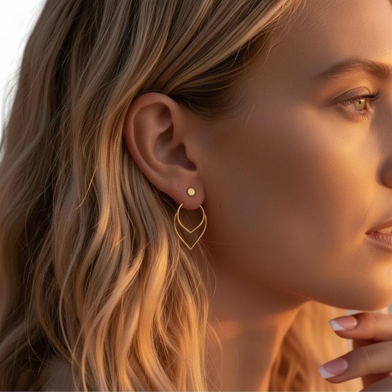 May include: Gold earrings featuring a small stud and a larger, leaf-shaped design. The earrings are worn on a person's ear, with the focus on the jewelry. The earrings are a bright gold color and appear to be made of metal. The earrings are a stylish accessory.