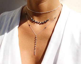 May include: A layered necklace set featuring three delicate chains. The top chain is a simple link design, the middle chain has a row of black beads, and the bottom chain has a vertical strand of black beads and a silver bar pendant. The jewelry is worn against a white top.