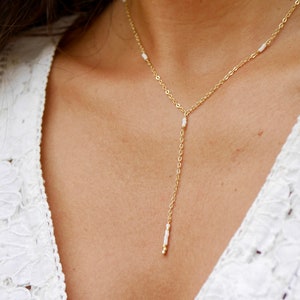 Puede incluir: Un collar de cadena dorada con pequeñas cuentas blancas. El collar es de estilo lariat, con una cadena larga y colgante.