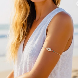 May include: A gold-colored arm cuff featuring a white seashell pendant. The arm cuff is adorned with small turquoise, pink, and gold beads. The jewelry is worn on the upper arm of a person wearing a white sleeveless top.