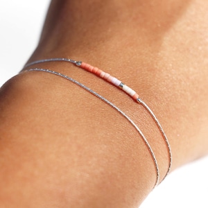May include: Two delicate silver bracelets on a person's wrist. The top bracelet features a row of small pink and white beads.