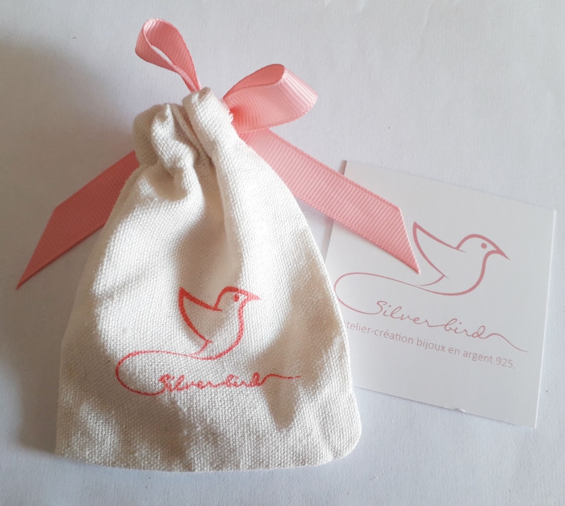 May include: A small, off-white drawstring bag with a pink ribbon and a matching tag. The bag features a red bird logo and the words "Silverbird". The tag also displays the Silverbird logo and text.