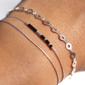 Pode incluir: Tr&ecirc;s pulseiras de prata empilhadas em um pulso. A pulseira superior tem uma corrente de pequenos c&iacute;rculos de prata. A pulseira do meio &eacute; uma corrente fina de prata. A pulseira inferior tem contas pretas.