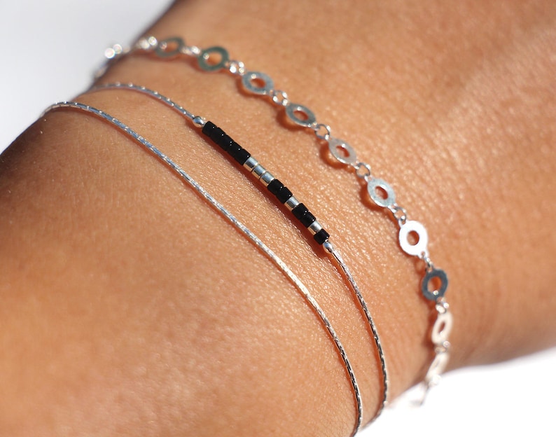 Pode incluir: Tr&ecirc;s pulseiras de prata empilhadas em um pulso. A pulseira superior tem uma corrente de pequenos c&iacute;rculos de prata. A pulseira do meio &eacute; uma corrente fina de prata. A pulseira inferior tem contas pretas.