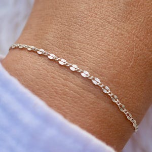 May include: A delicate silver chain bracelet with a delicate, textured, leaf-like pattern. The bracelet is shown on a person's wrist.