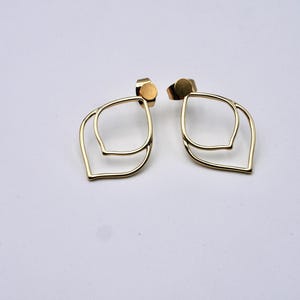May include: A pair of gold-colored, open-frame earrings. Each earring features a double-layered, leaf-like design. The earrings have a post and backing for secure wear. The background is a plain white surface.