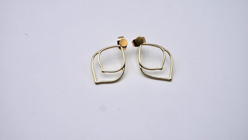 May include: A pair of gold-colored, open-frame earrings. Each earring features a double-layered, leaf-like design. The earrings have a post and backing for secure wear. The background is a plain white surface.