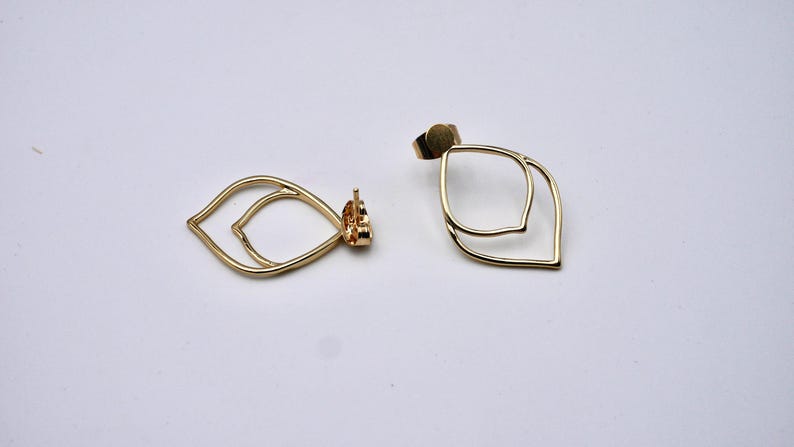 May include: A pair of gold-colored, open-frame earrings. Each earring features a double leaf design. The earrings are displayed on a white surface, showcasing their delicate, minimalist style. The earrings have post backs.