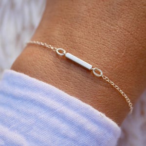 Pode incluir: Uma delicada pulseira de prata com um pequeno pendente retangular. A pulseira é presa a uma corrente fina e é usada em um pulso com uma manga branca.