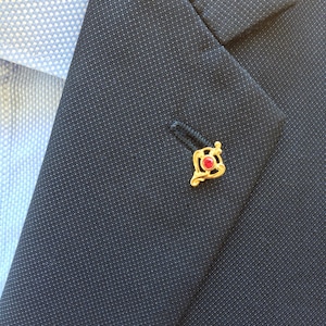 May include: A gold lapel pin with a red gemstone in the centre. The pin has a decorative design with intricate details.