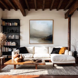 Pode incluir: Uma sala de estar com um grande sofá seccional branco, uma mesa de centro de madeira e uma grande pintura abstrata. O quarto tem uma paleta de cores neutras, luz natural e vigas de madeira no teto.