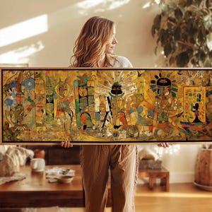 May include: A rectangular framed artwork with a detailed, colourful illustration. The artwork features figures and symbols in shades of yellow, blue, red, and green. The frame is a light brown colour. The artwork is held by a person.